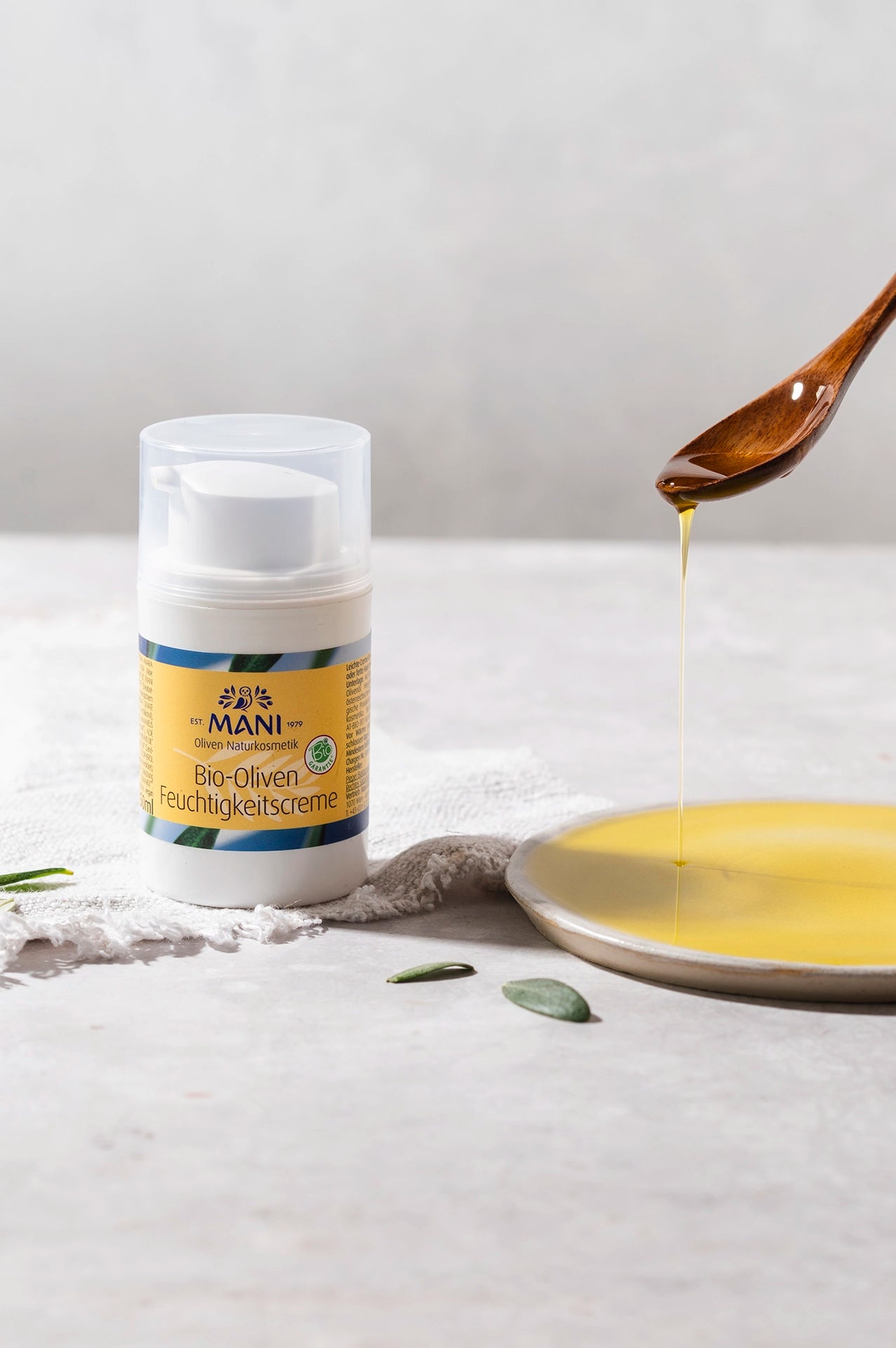 MANI Bio Oliven Feuchtigkeitscreme neben Olivenöl, das vom Löffel auf einen Teller fließt