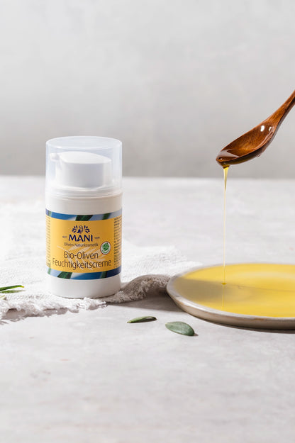 MANI Bio Oliven Feuchtigkeitscreme neben Olivenöl, das vom Löffel auf einen Teller fließt