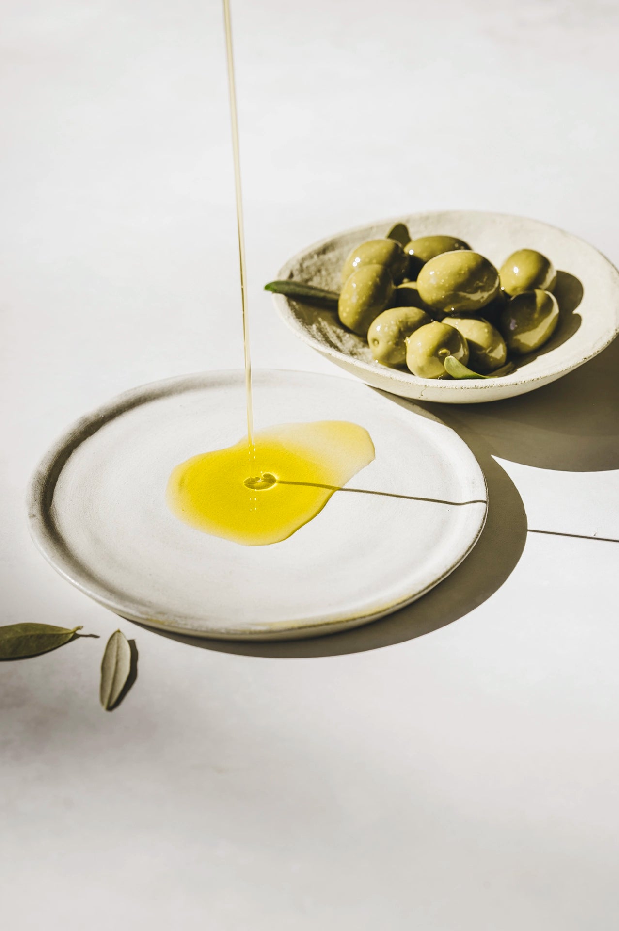 MANI Olivenöl fließt auf weißen Teller, Oliven in Schälchen im Hintergrund